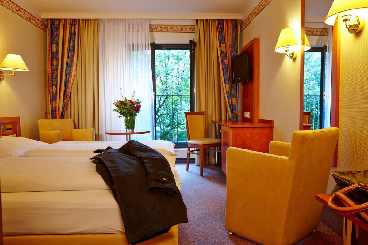 A hotel room with two beds, a desk, a chair, a TV, and a balcony with curtains.