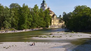 A riverside view with a historic building in the background, a pebbled beach, and trees lining the riverbank. - Hotel Concorde München (Munich)