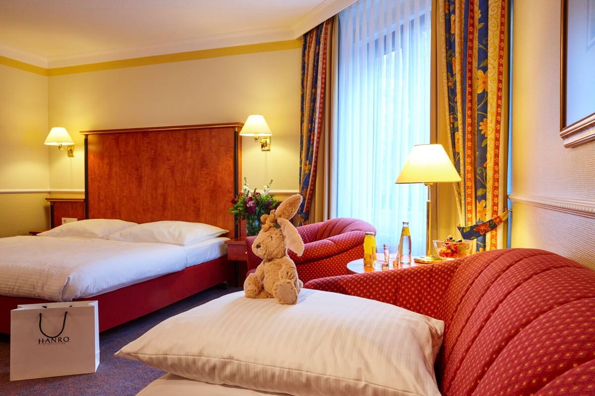 A hotel room with two beds, a red sofa, a teddy bear, a lamp, and a window with curtains.