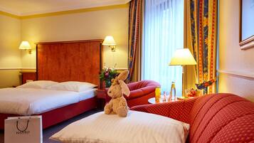 A hotel room with two beds, a red sofa, a teddy bear, a lamp, and a window with curtains.