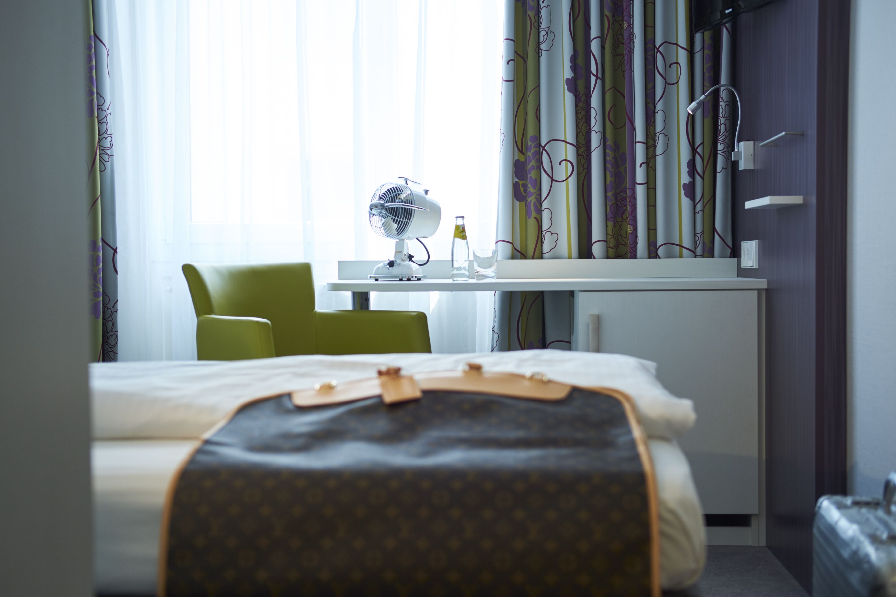 A hotel room with a bed, a desk with a fan, a chair, and a suitcase.