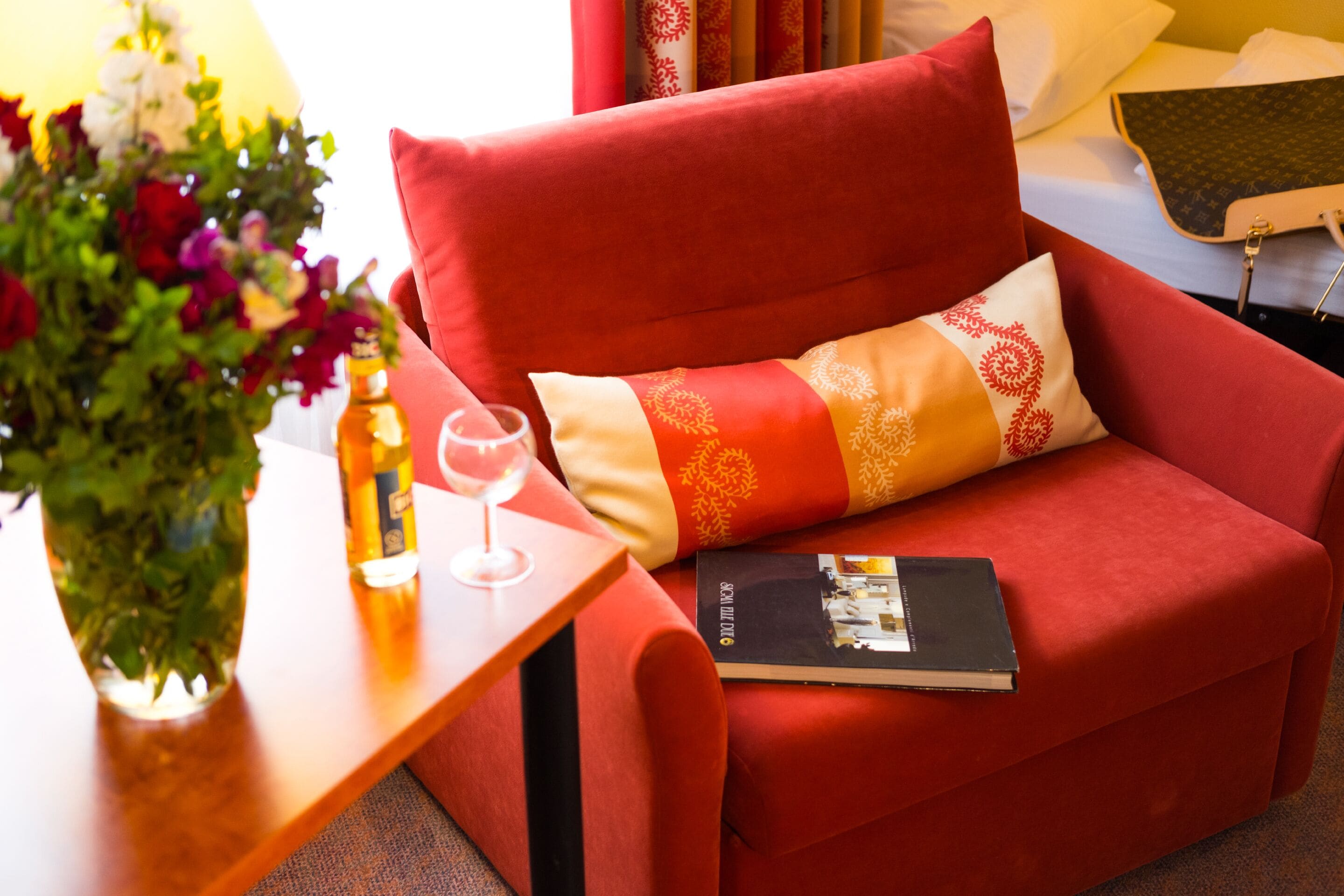 A red sofa with a patterned pillow, a book, a glass of wine, and a vase of flowers on a wooden side table.