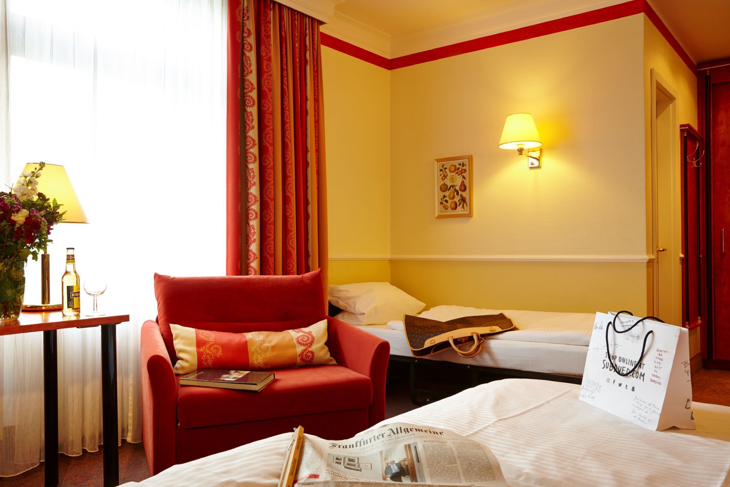 Habitación de hotel con un sillón rojo, una cama con edredón blanco, una mesita de noche con lámpara y una lámpara de pared.