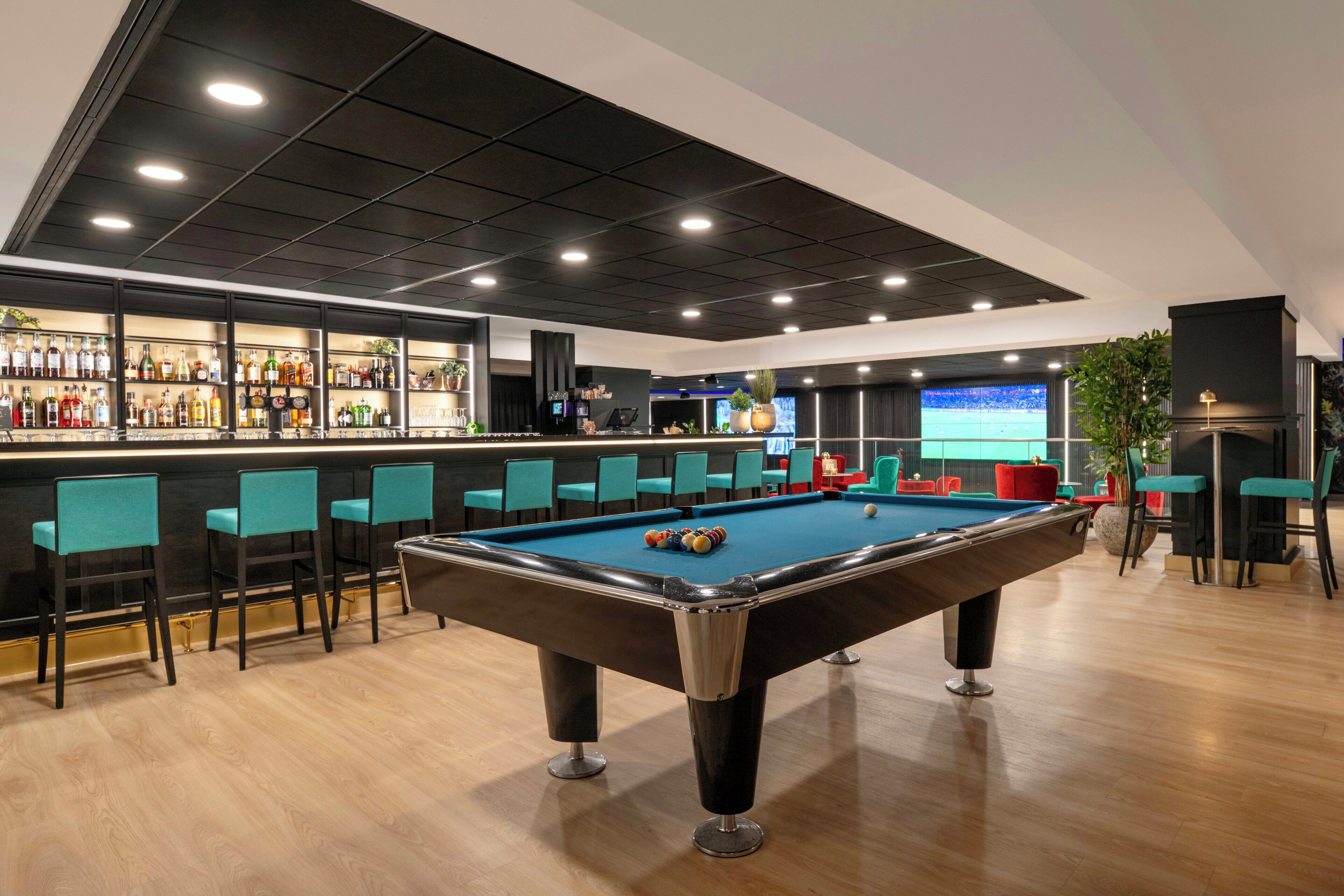 A pool table with chairs, a bar area with bottles, and a TV screen in a modern indoor setting.