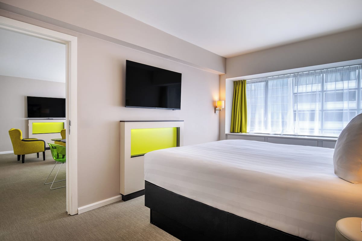 A hotel room with a bed, a TV mounted on the wall, a desk with a chair, and a window with curtains.