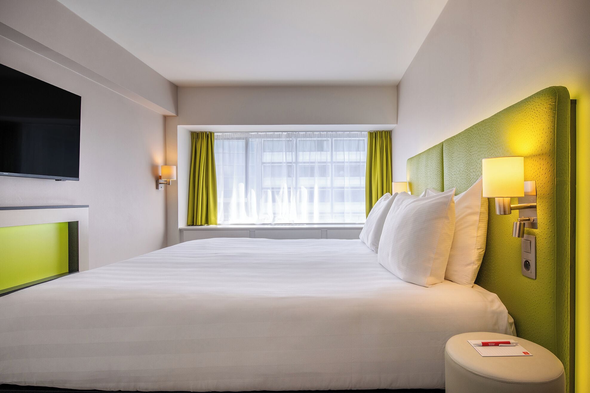 A hotel room with a large bed, a flat-screen TV mounted on the wall, a window with green curtains, and a green accent wall.