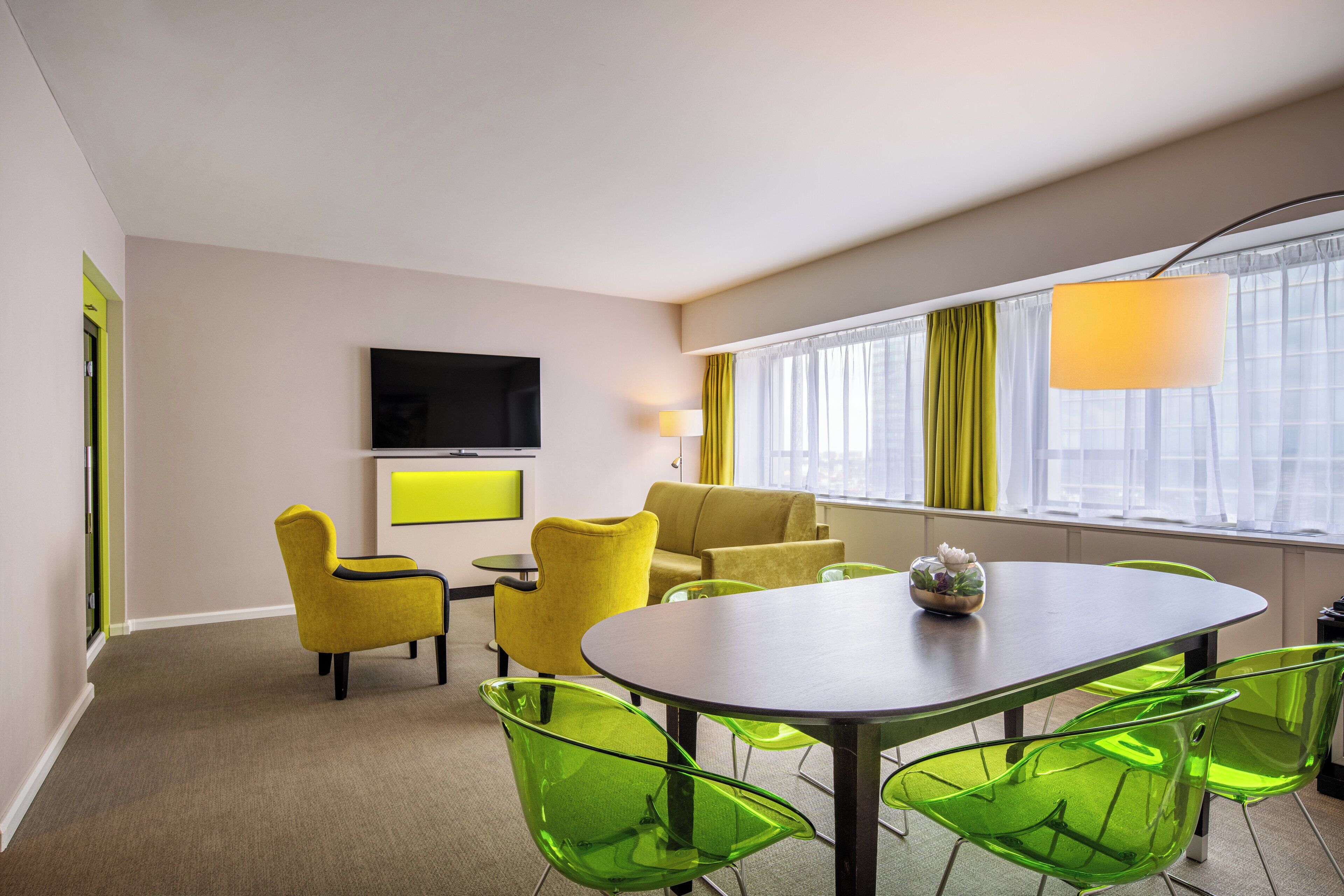 A modern hotel room with a dining area, a flat-screen TV mounted on the wall, and a large window with curtains.