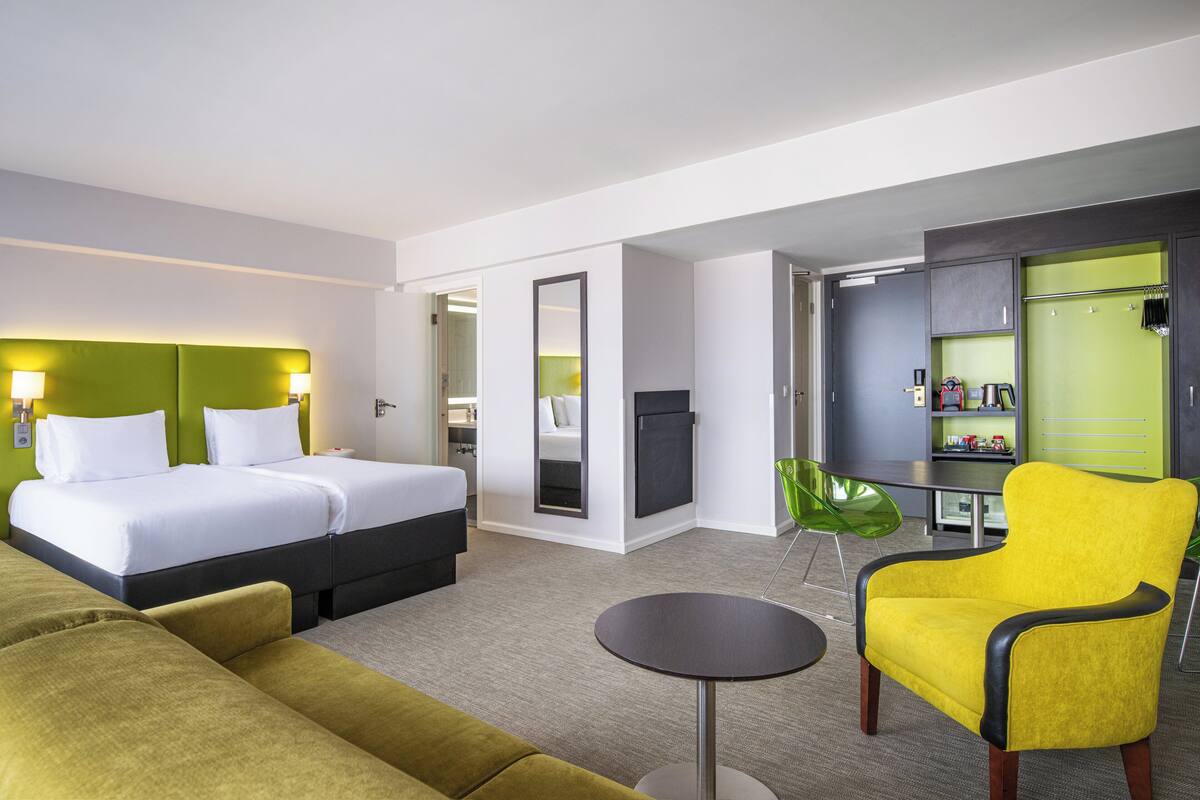 A hotel room with a bed, a green sofa, a yellow armchair, and a small table.