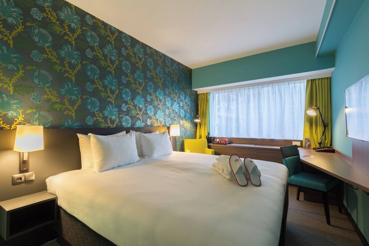 A hotel room with a large bed, a desk, and a chair. There is a window with curtains, a lamp, and a nightstand.