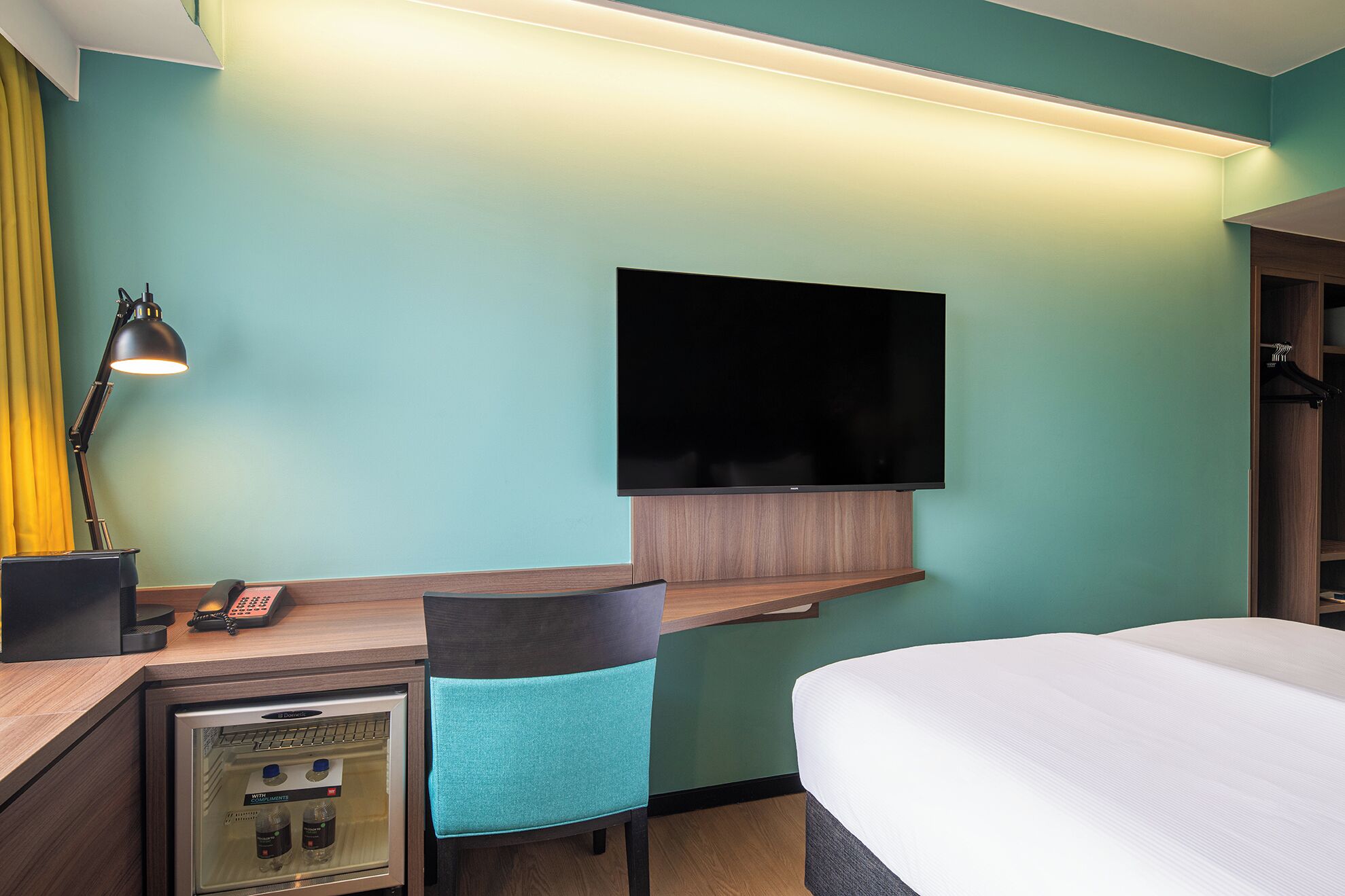 Habitación de hotel con cama, escritorio con silla, televisor montado en la pared, minibar y lámpara.