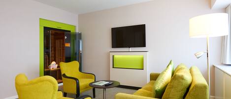 A modern hotel room with yellow armchairs, a small round table, a flat-screen TV, and a lamp.