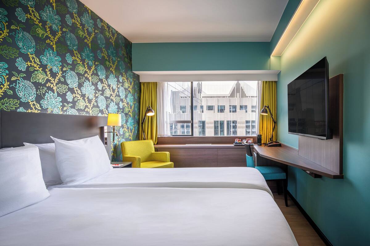 A hotel room with a large bed, a desk, a chair, a TV, and a window with a city view.