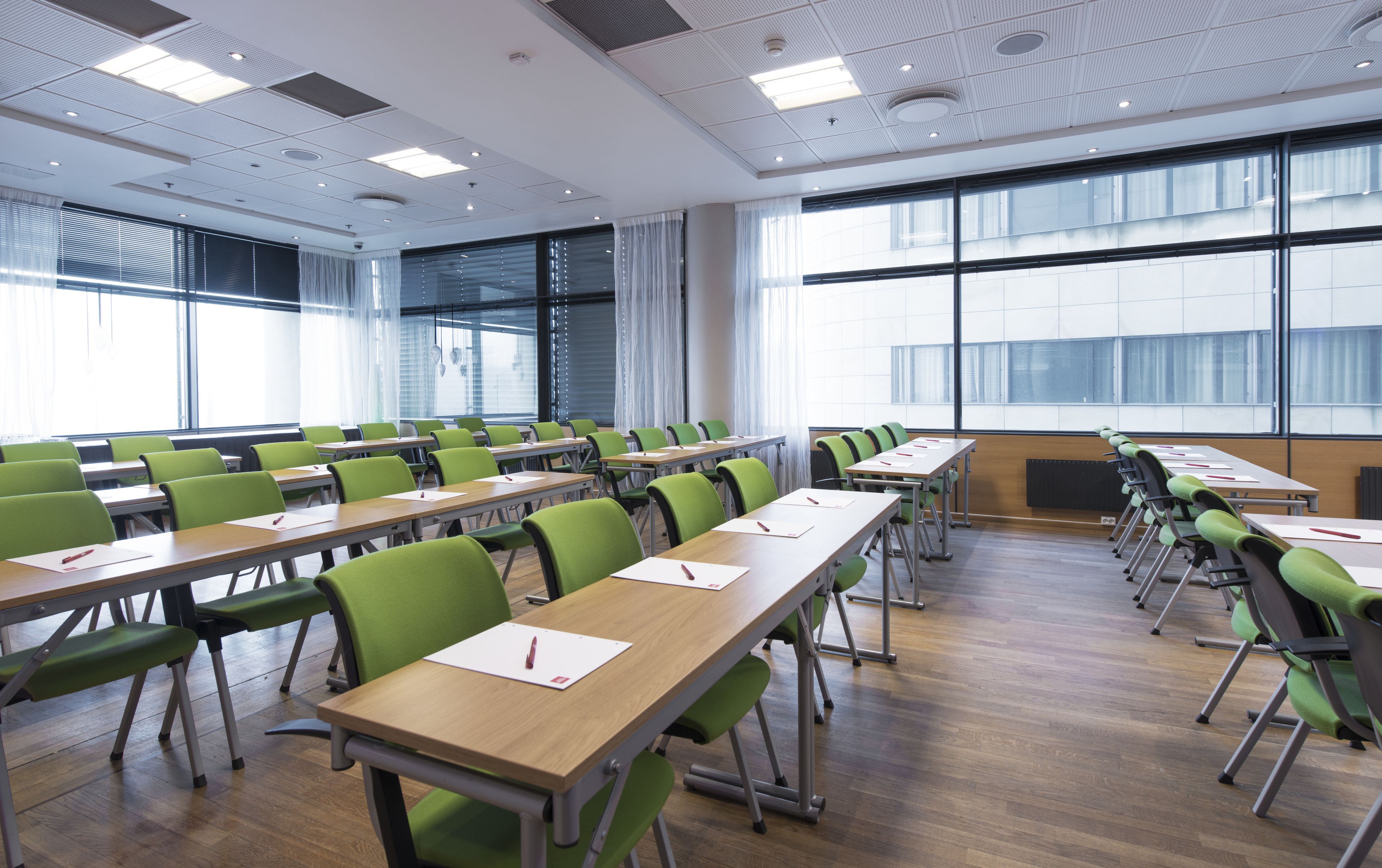 A conference room with rows of green chairs and wooden tables, large windows, and a modern ceiling design.