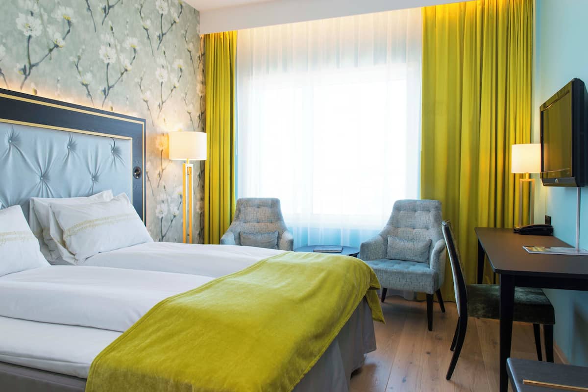 A hotel room with a large bed, two armchairs, a desk, a television, and a window with curtains.