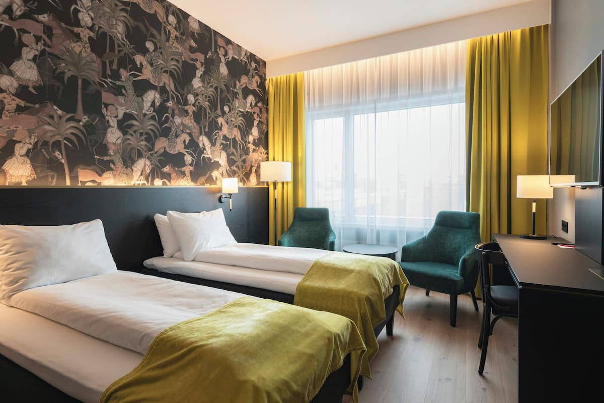 Habitación de hotel con dos camas, un escritorio, dos sillas, un ventanal con cortinas y un mural en la pared.