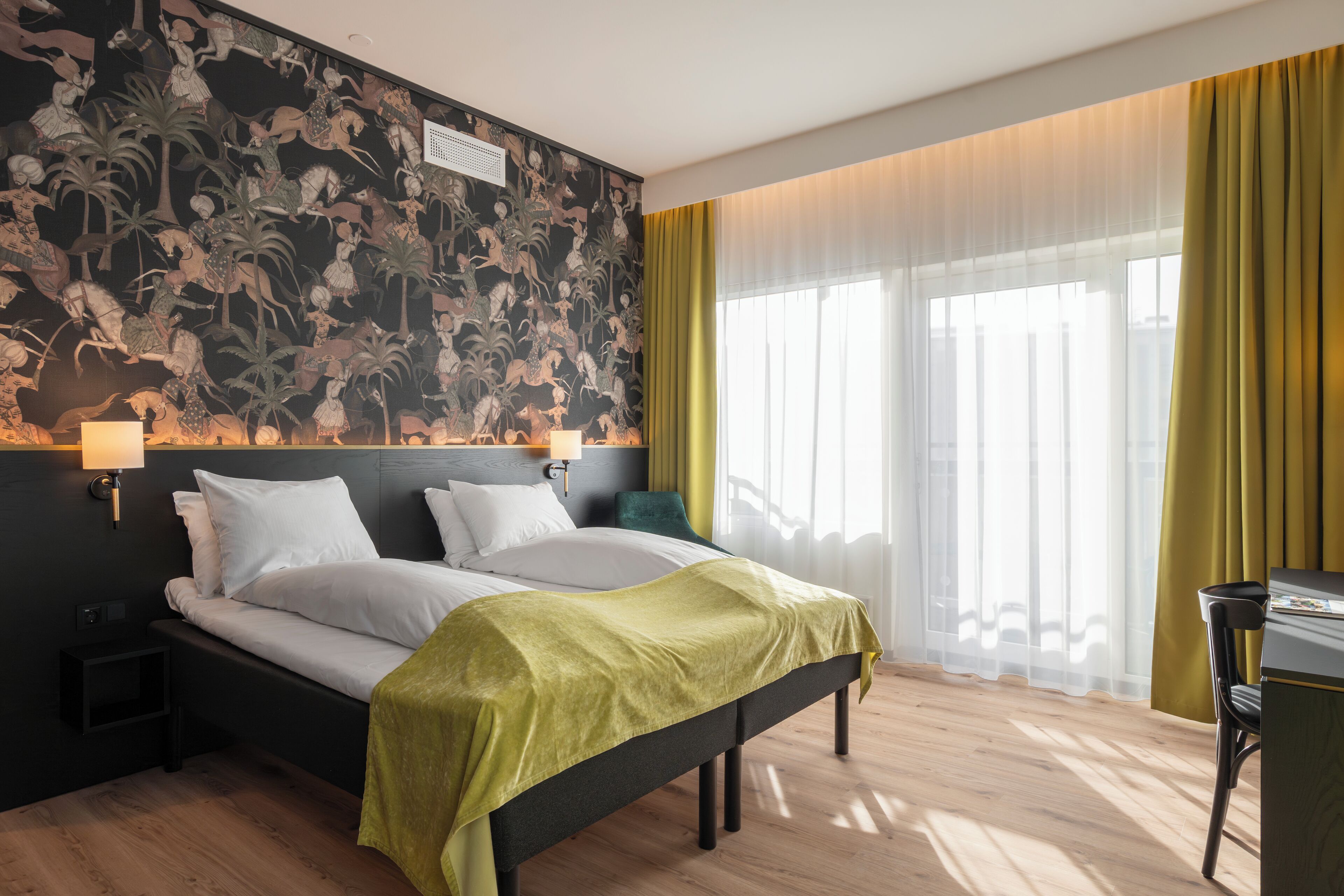 Habitación de hotel con cama, escritorio, silla, mesita de noche, pared con mural y ventana con cortinas.