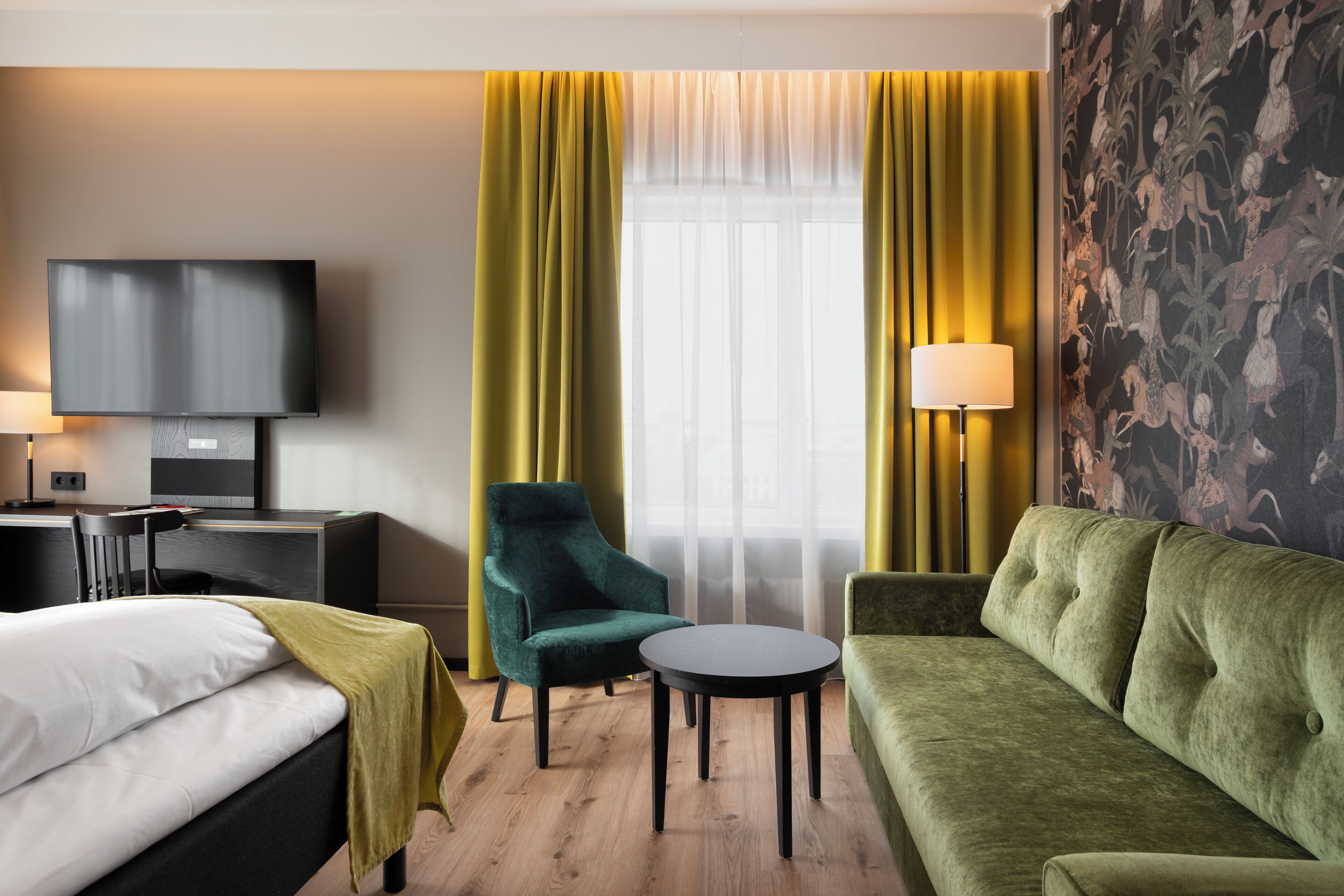 Habitación de hotel con cama, un sillón verde, un sofá, una mesita redonda, un televisor y una lámpara.
