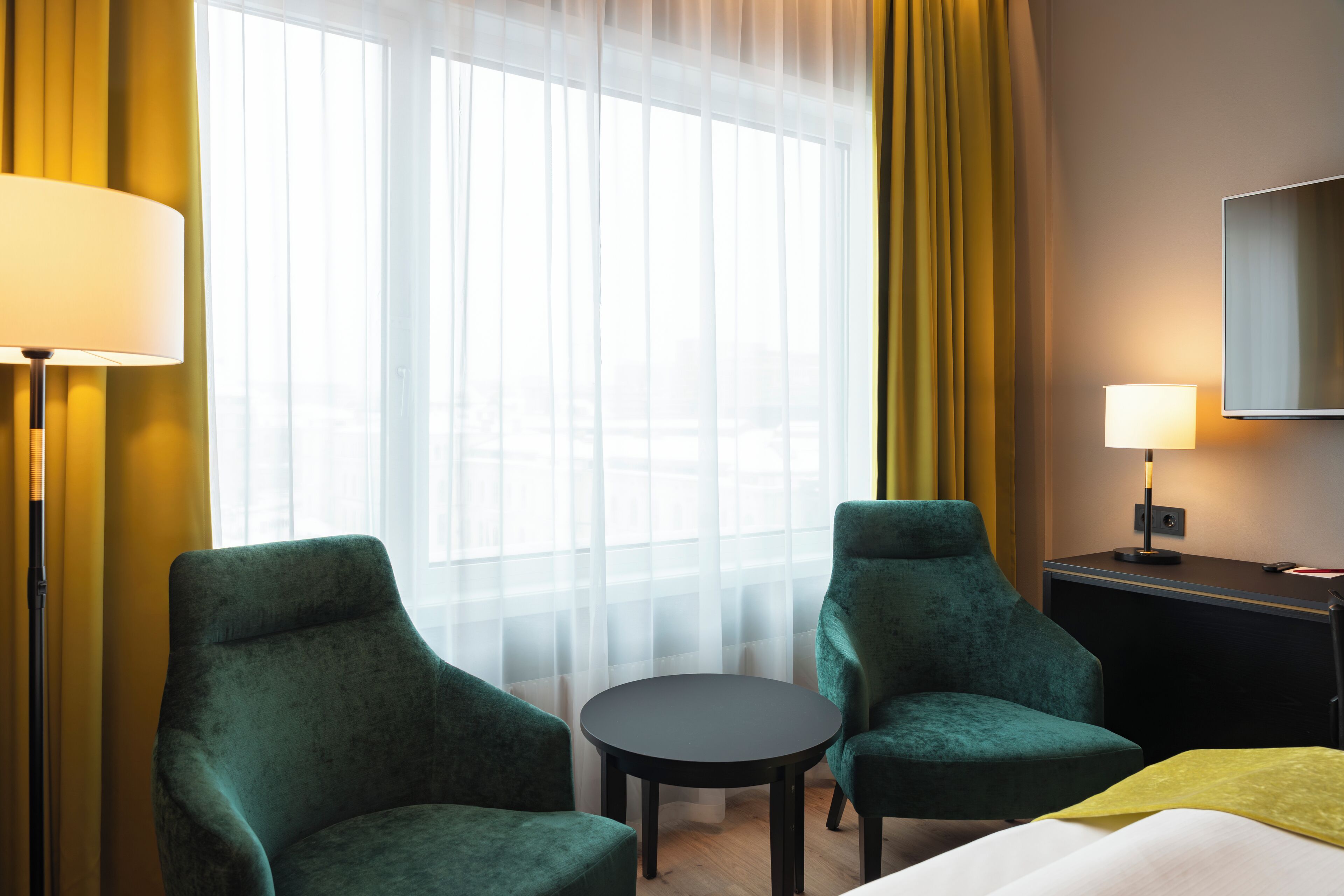 A hotel room with two green armchairs, a small round table, a bed with a yellow blanket, a TV on the wall, and a lamp on the nightstand.