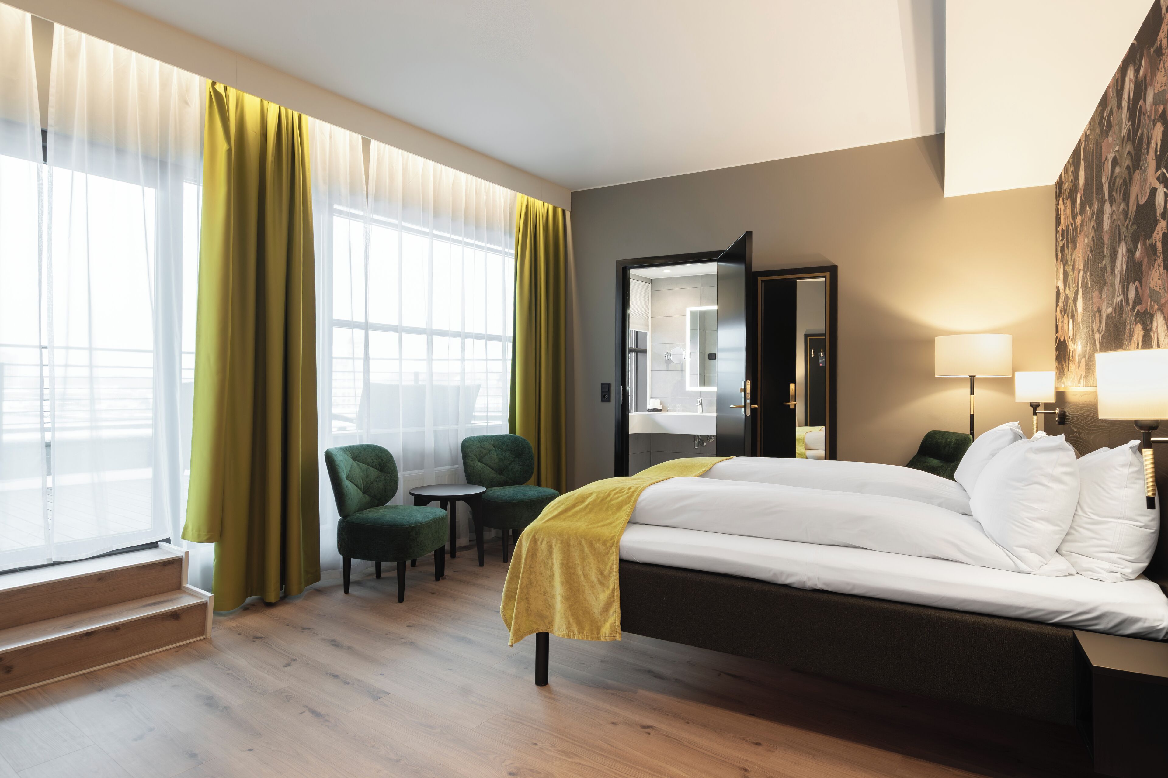 Habitación de hotel con cama, una silla, una mesita, un espejo y una ventana con cortinas.