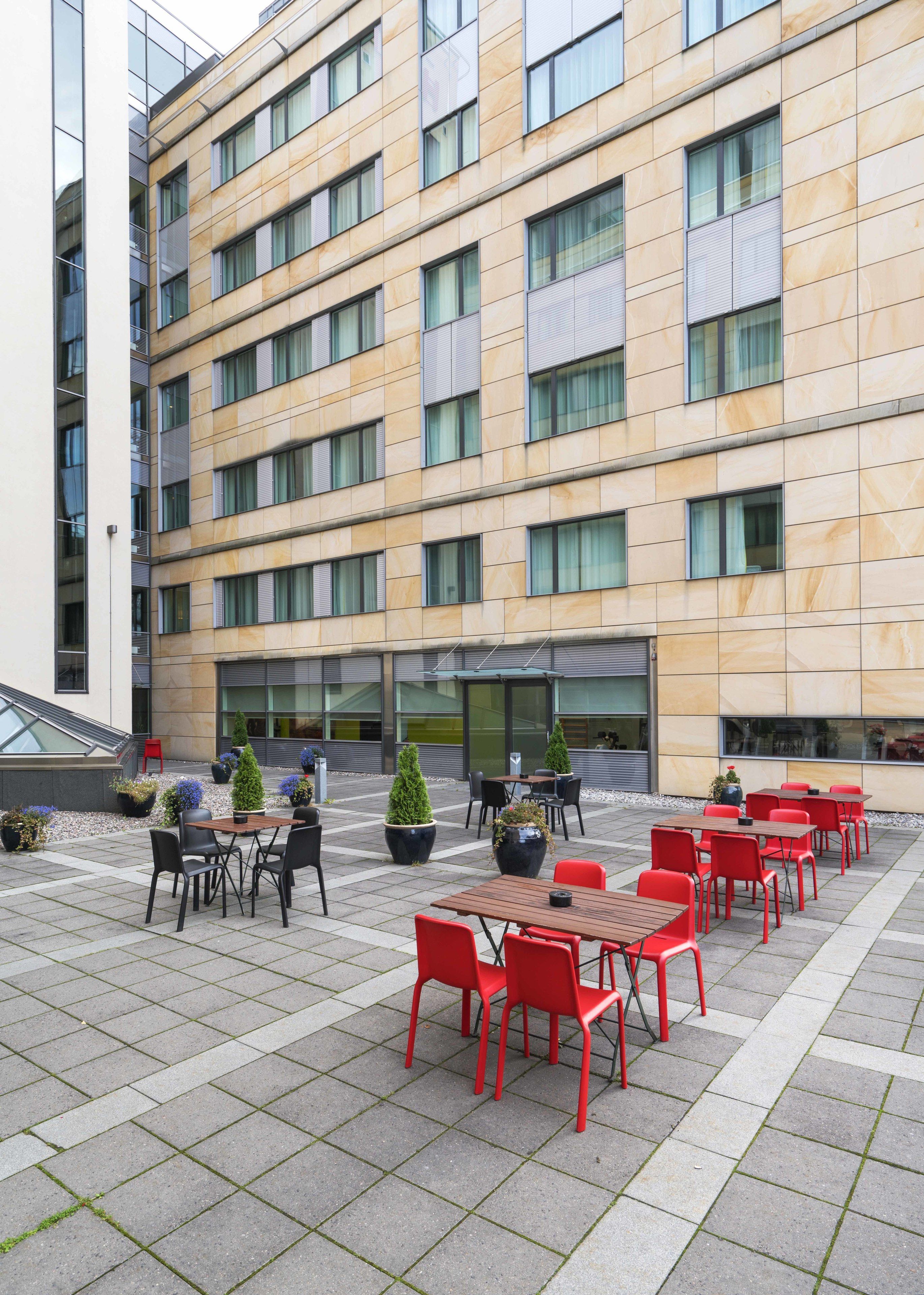 A modern building with a courtyard featuring outdoor seating with red chairs and tables, potted plants, and a staircase leading to an upper level.
