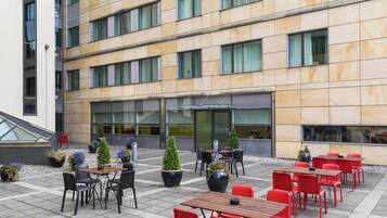 A modern building with a courtyard featuring outdoor seating with red chairs and tables, potted plants, and a staircase leading to an upper level.