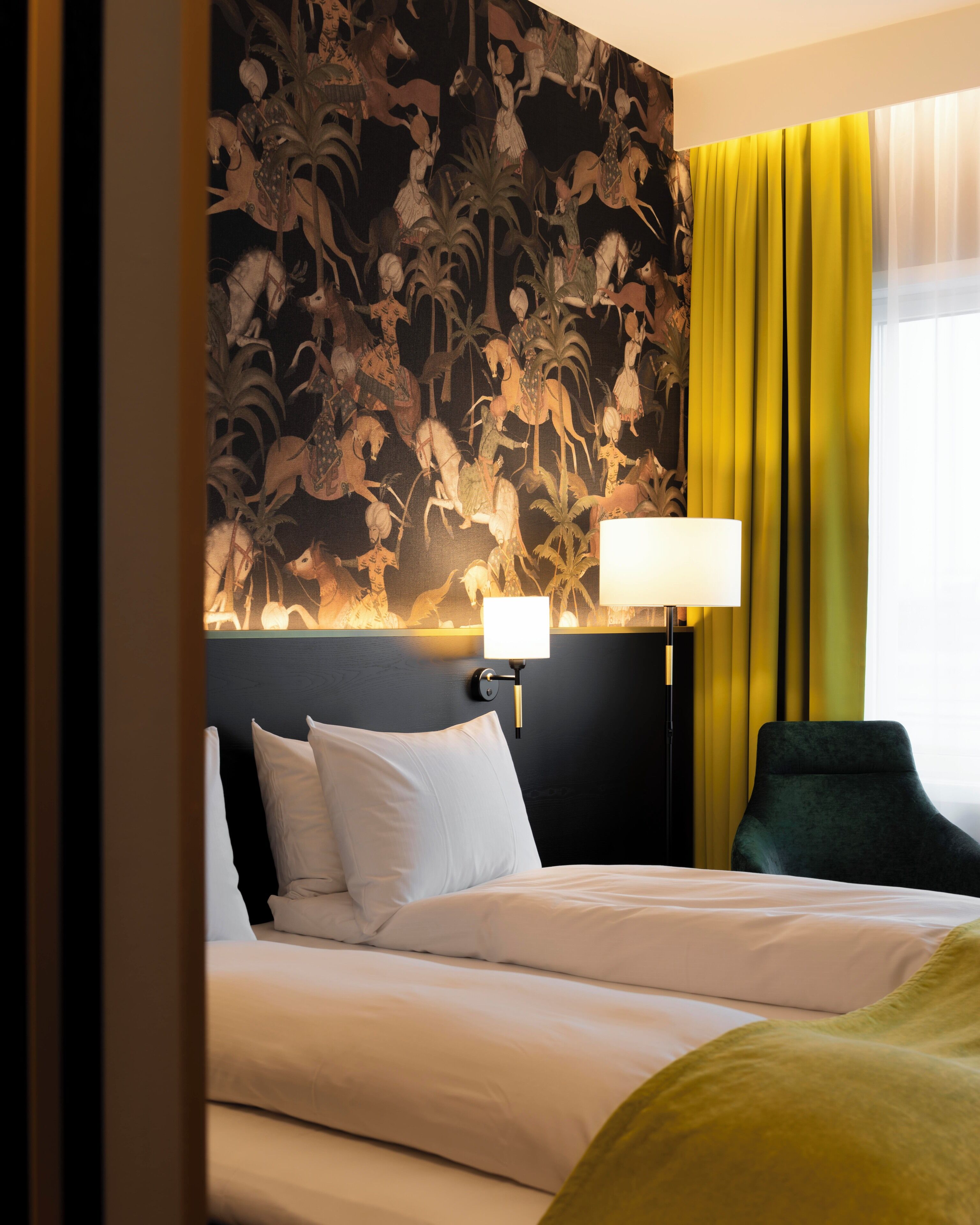 A hotel room with a bed, a nightstand with a lamp, a chair, and a wall with a floral pattern.