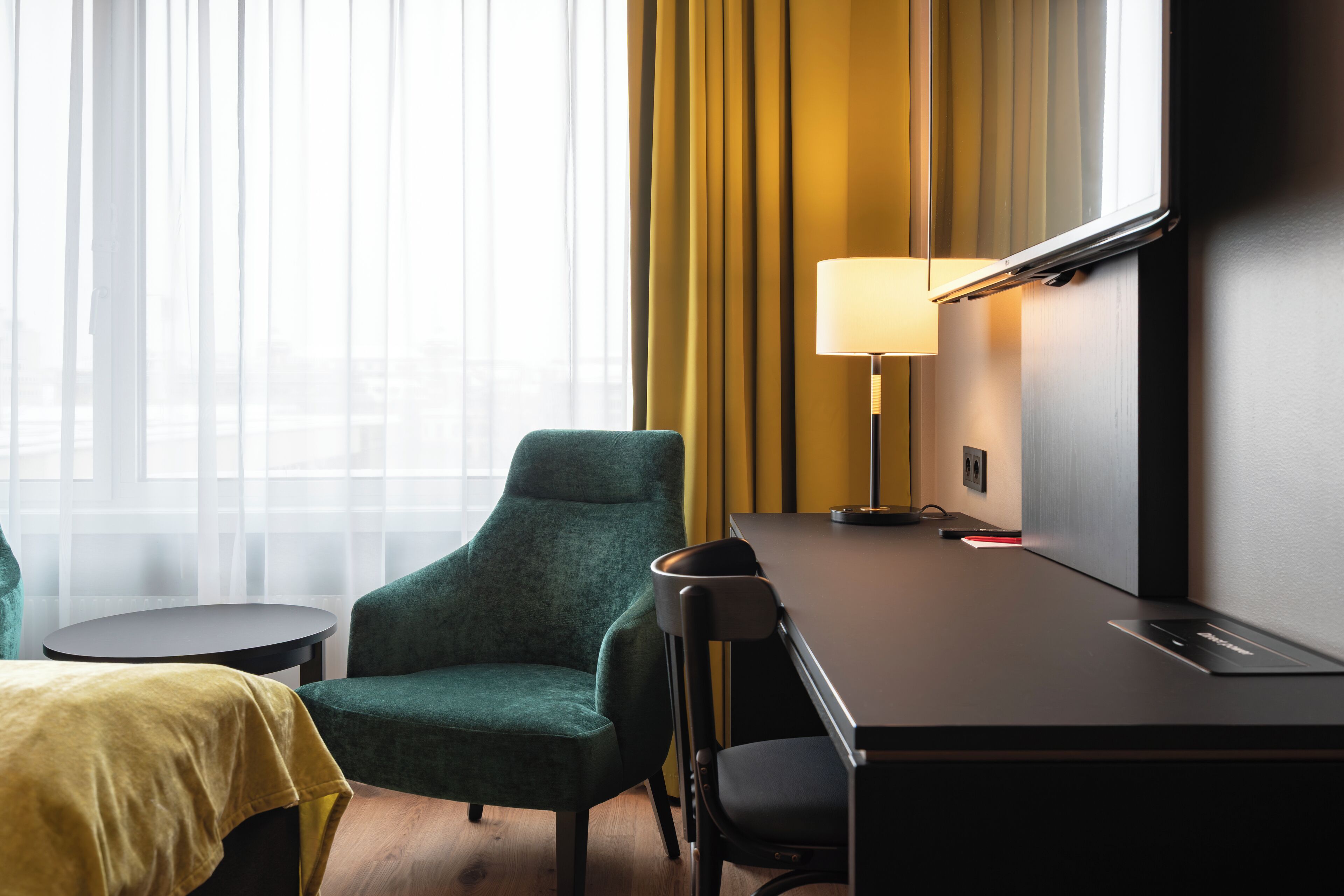 A hotel room with a desk, a green armchair, a small round table, a lamp, and a view through sheer curtains.