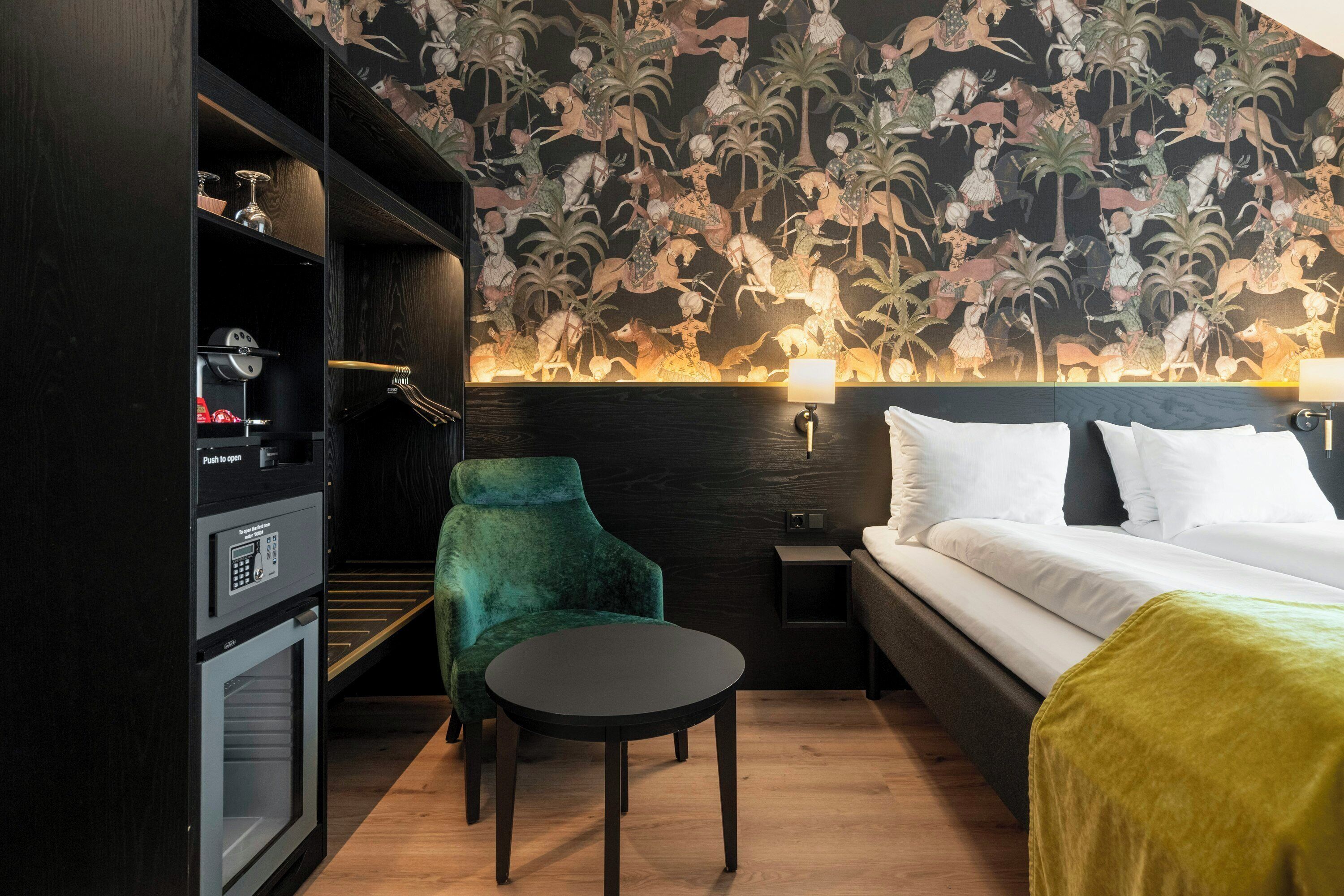 A hotel room with a bed, a green chair, a small table, a minibar, and a wall with a tropical mural.