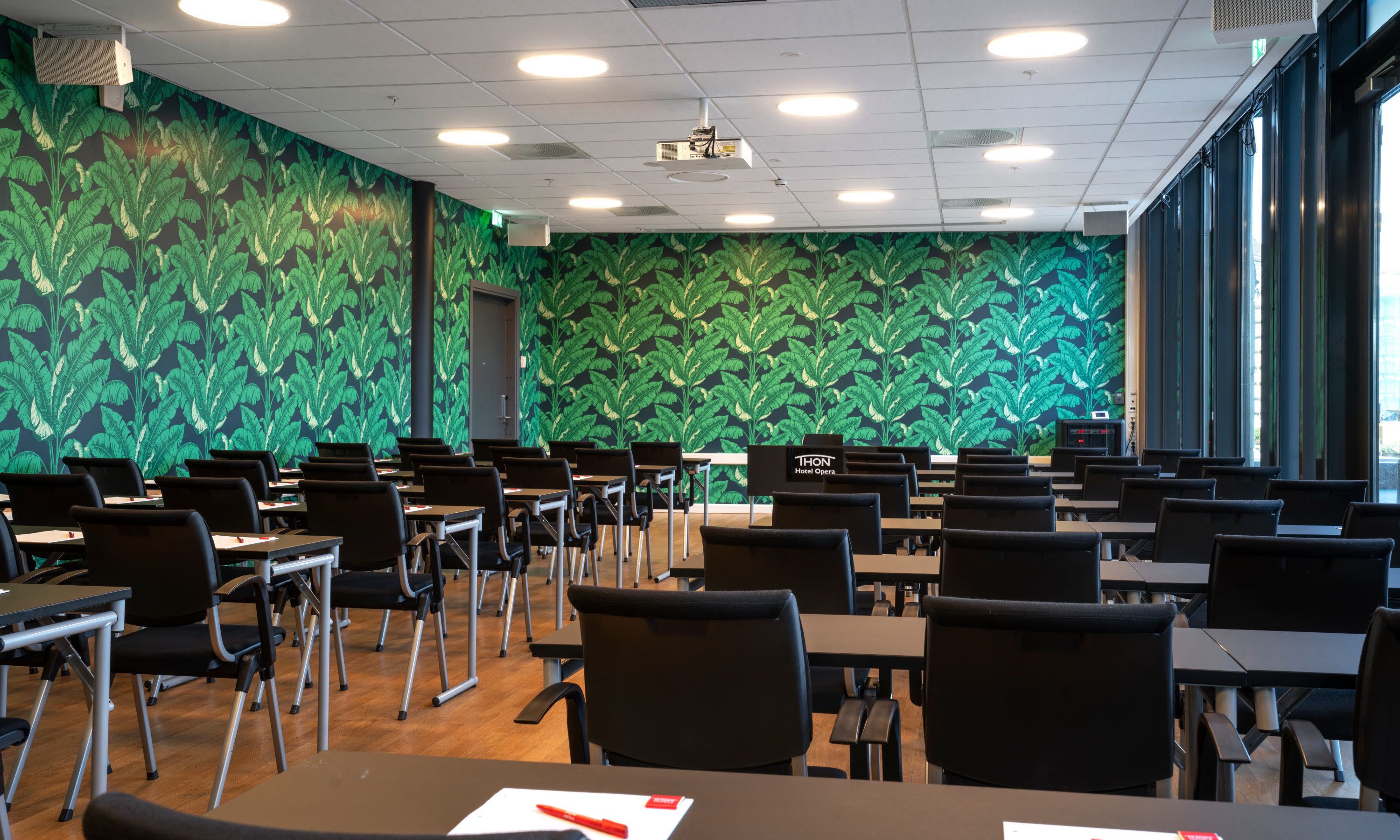 Un salón de conferencias con pared estampada con motivos de hojas verdes, varias mesas y sillas.