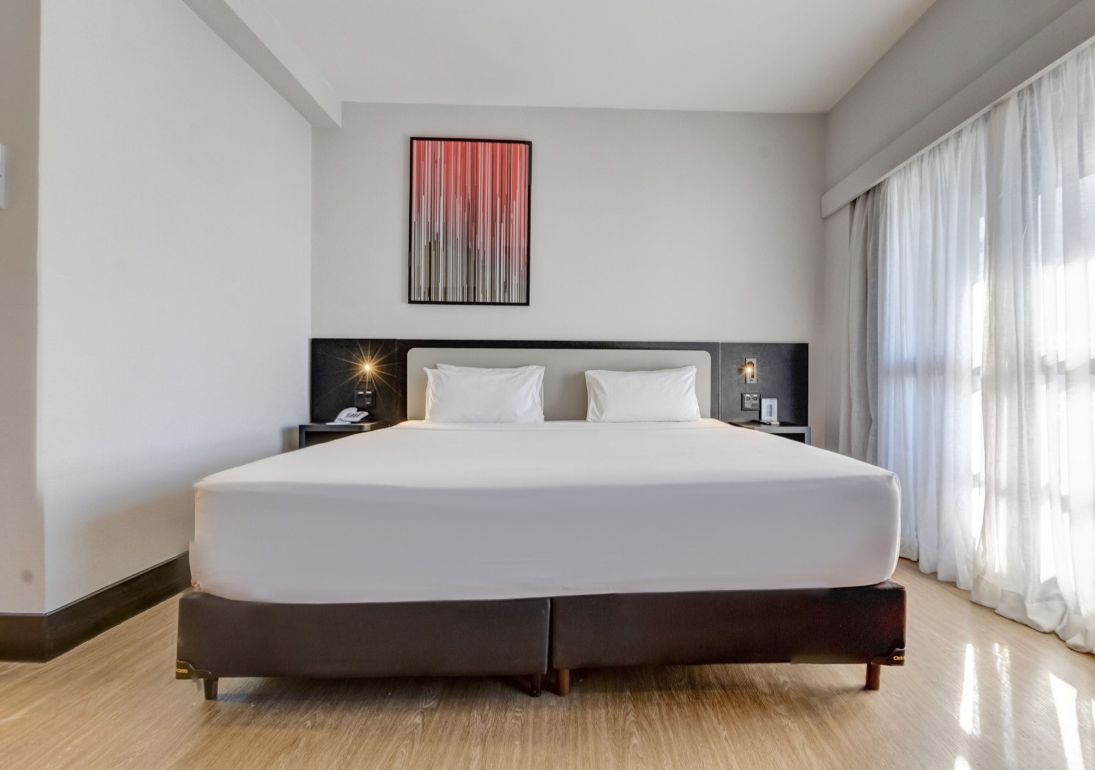 A large bed with a dark frame, a headboard with a light fixture, a bedside table with a lamp, and a large window with curtains.