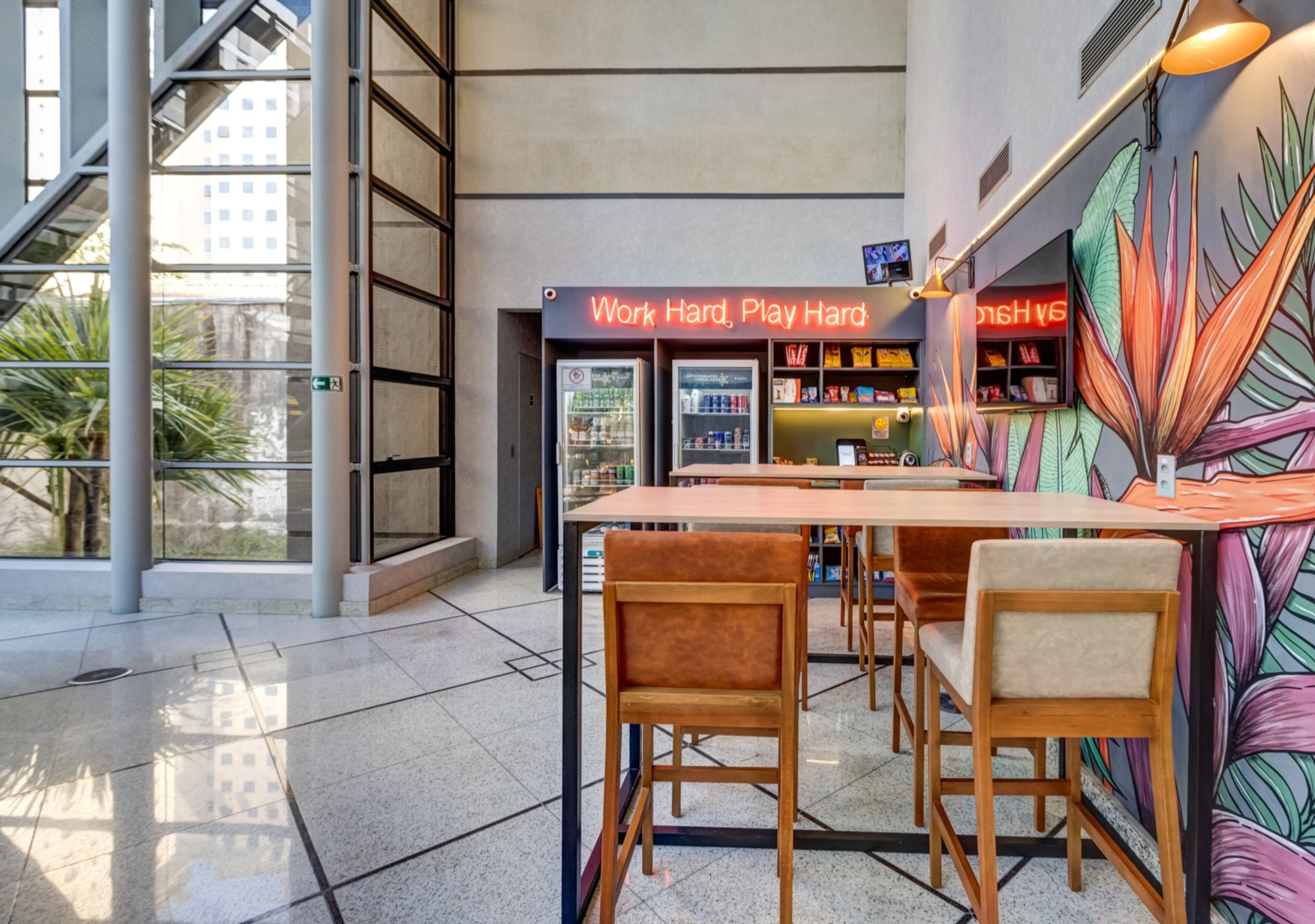 A modern indoor space with a long table and chairs, a neon sign reading 'Work Hard, Play Hard', and a colorful mural on the wall.