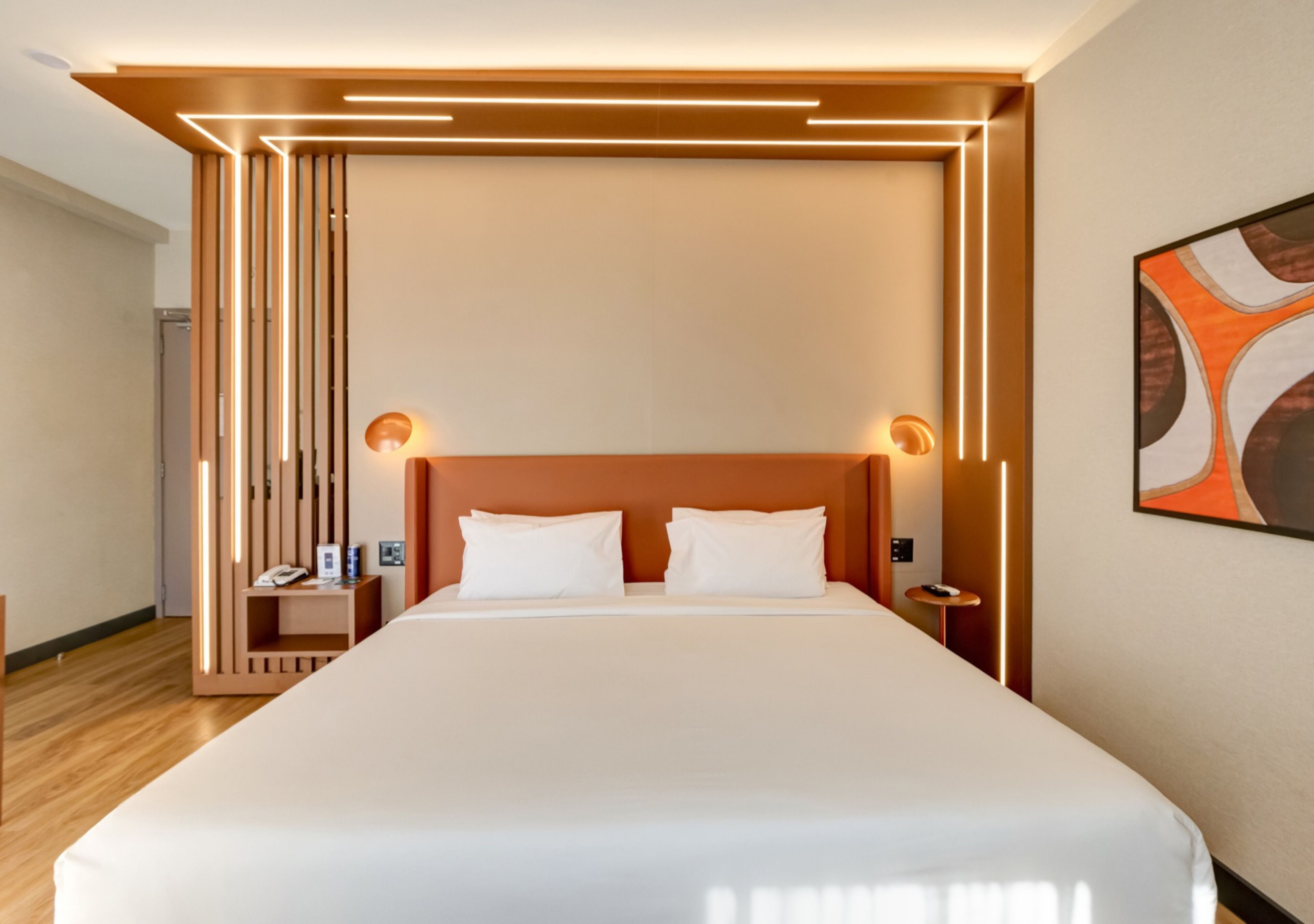 A modern hotel room with a large bed, wooden headboard, bedside lamps, and a wall-mounted artwork.