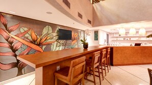 Zona de bar moderna con una barra alargada, varios taburetes, un mural con plantas tropicales, un televisor de pantalla plana y una escalera al fondo.