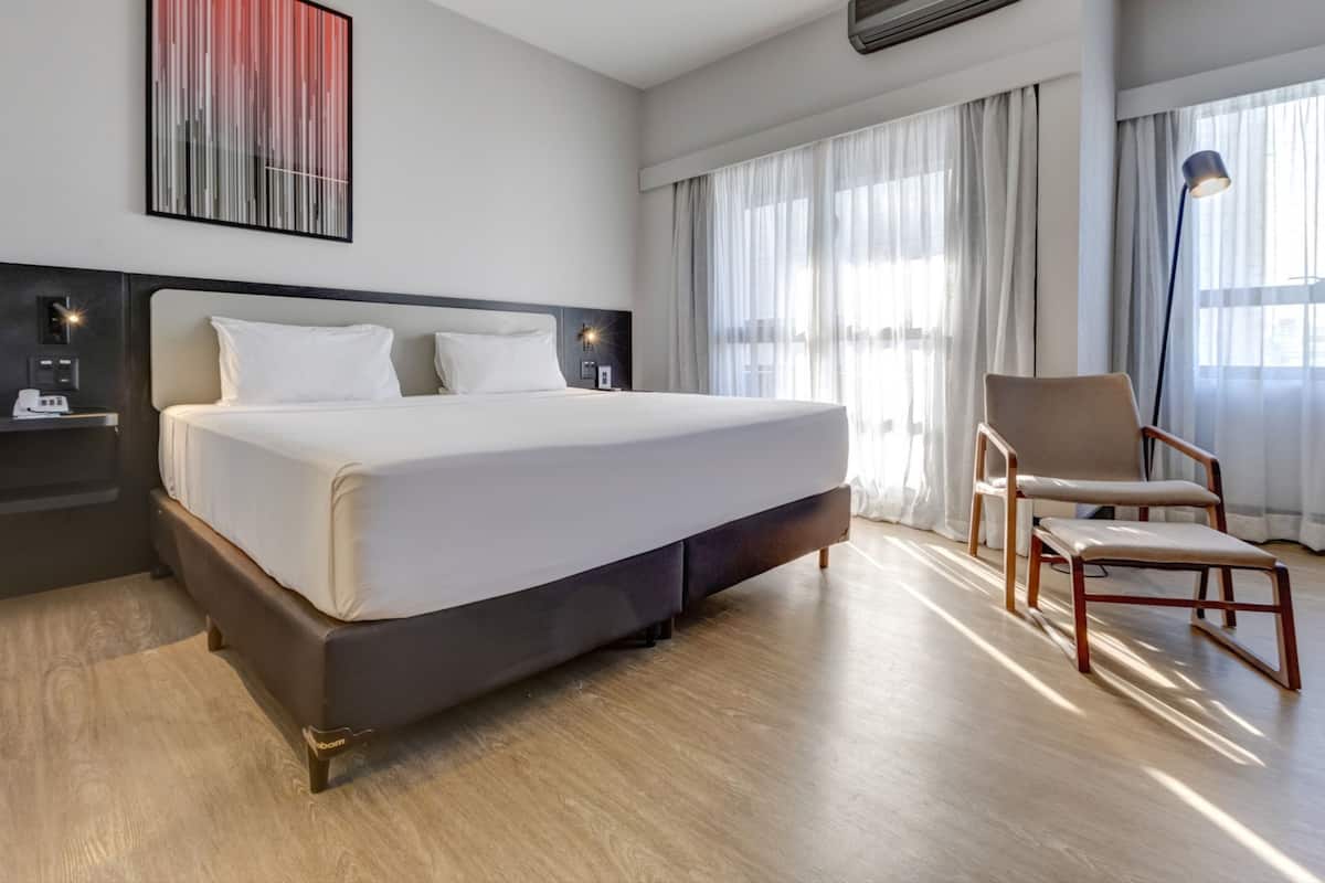 A hotel room with a large bed, a chair, a bedside table with a lamp, and a wall-mounted air conditioner.