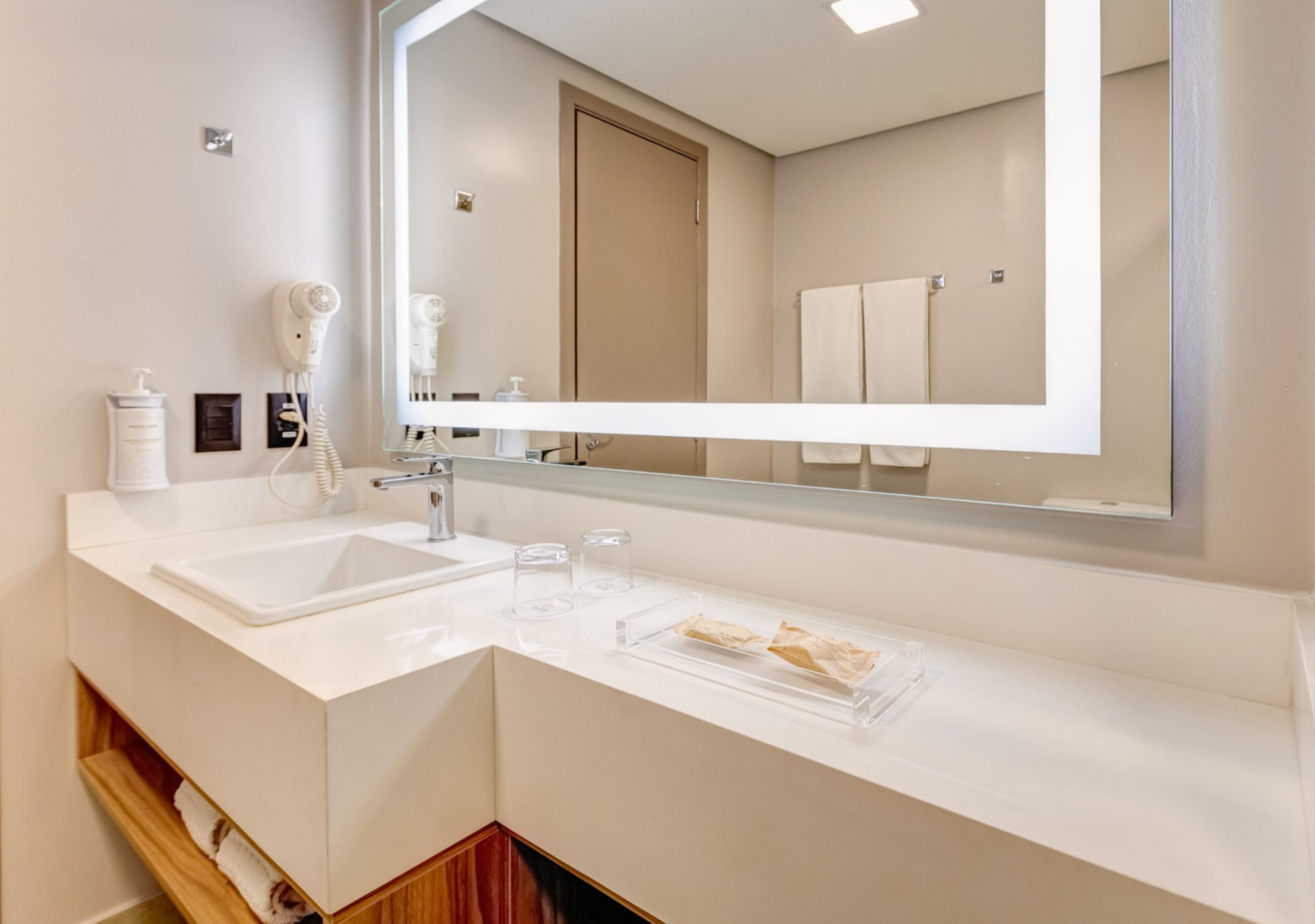Een moderne badkamer met een witte wastafel, een grote spiegel en een haardroger aan de muur.