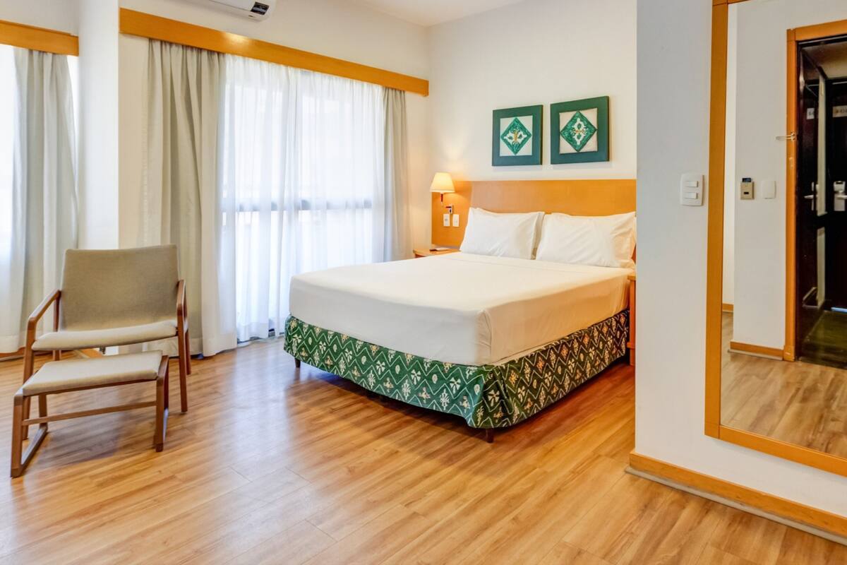 A hotel room with a bed, a chair, a nightstand, a lamp, and two framed pictures on the wall.