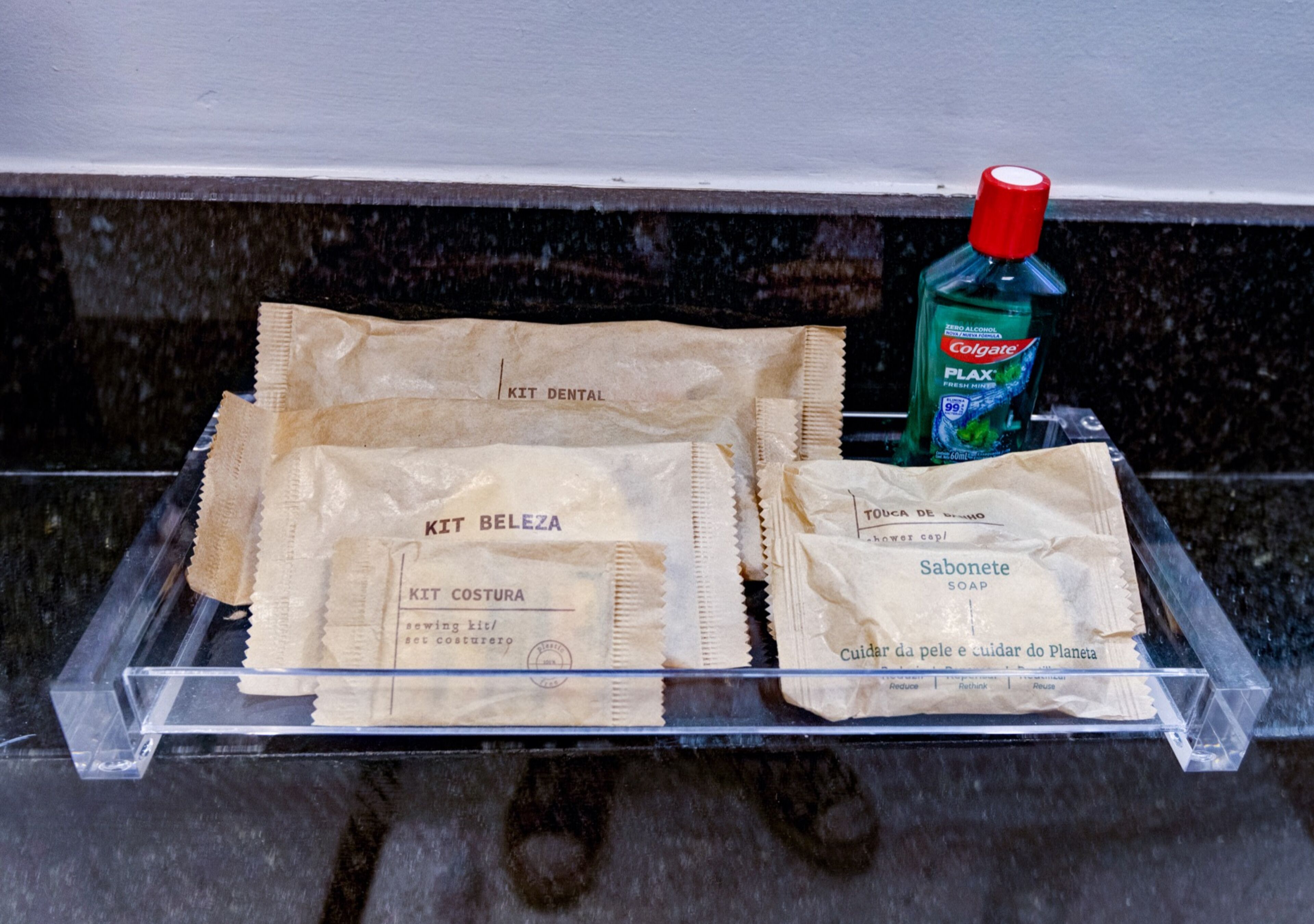 Ein Badezimmer mit einer klaren Kunststoffschale, die Toilettenartikel enthält, darunter eine Tube Colgate Zahnpasta und zwei Beutel mit der Aufschrift 'KIT BELEZA' und 'KIT COSTURA'.