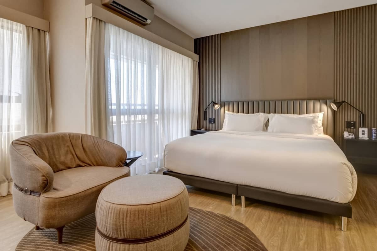 A modern hotel room with a large bed, a sofa, an ottoman, a nightstand, and a window with curtains.