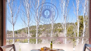 Una mesa de desayuno con un plato de fruta, jugo y pan, además de una botella de aceite de oliva, con vistas a árboles y colinas a través de una ventana.