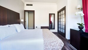 Habitación de hotel con una cama grande, una mesita de noche, un espejo, una ventana con vistas y una puerta que da a otra habitación.