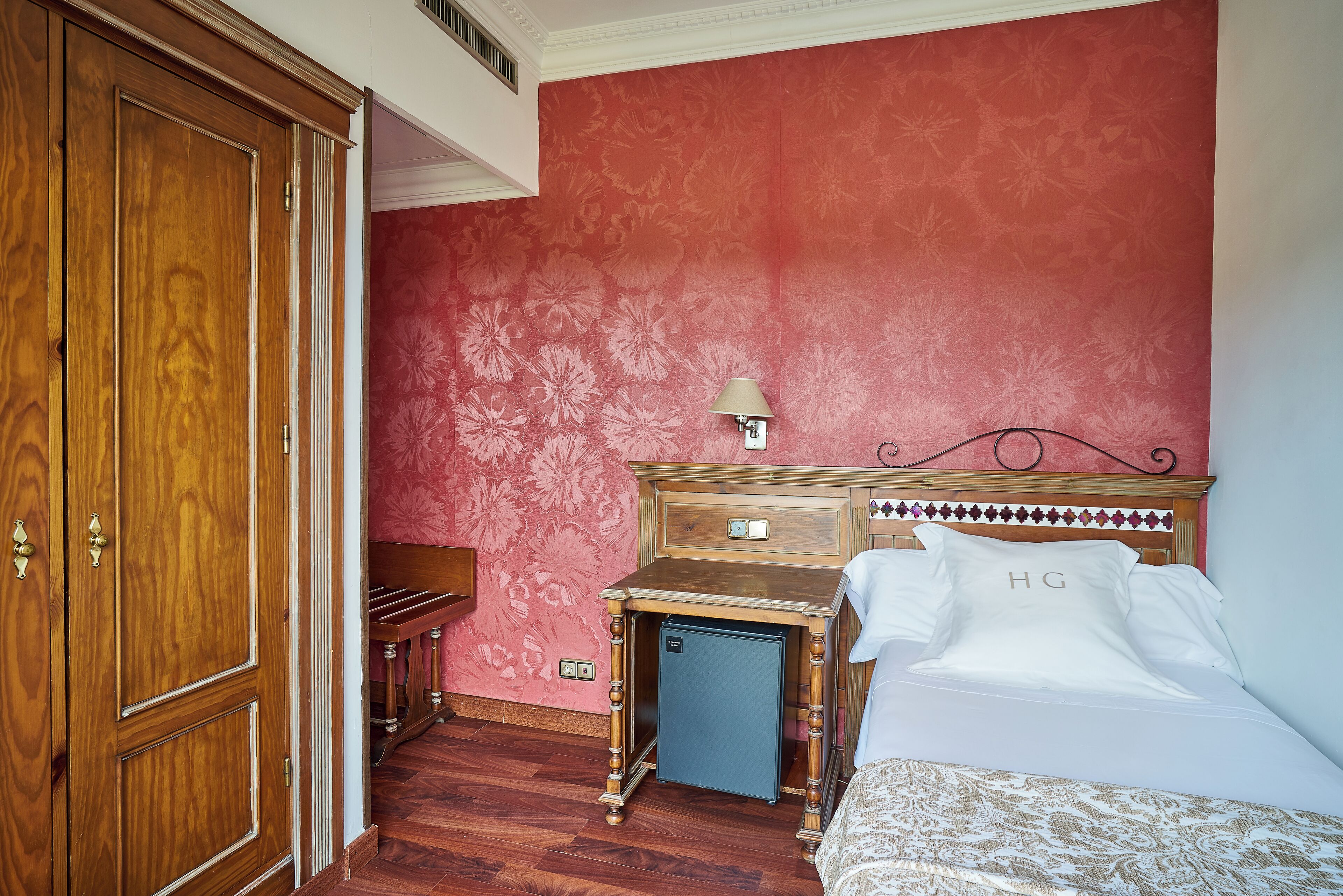 Quarto com guarda-roupa de madeira, uma pequena escrivaninha com luminária e uma cama com travesseiro branco com a marca 'HG'.