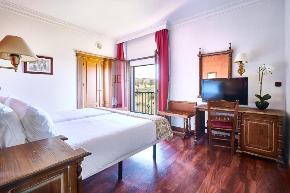 A hotel room with a large bed, a desk with a chair, a television, and a balcony with red curtains.