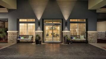 A building entrance with a glass door, two benches with cushions, and potted plants.