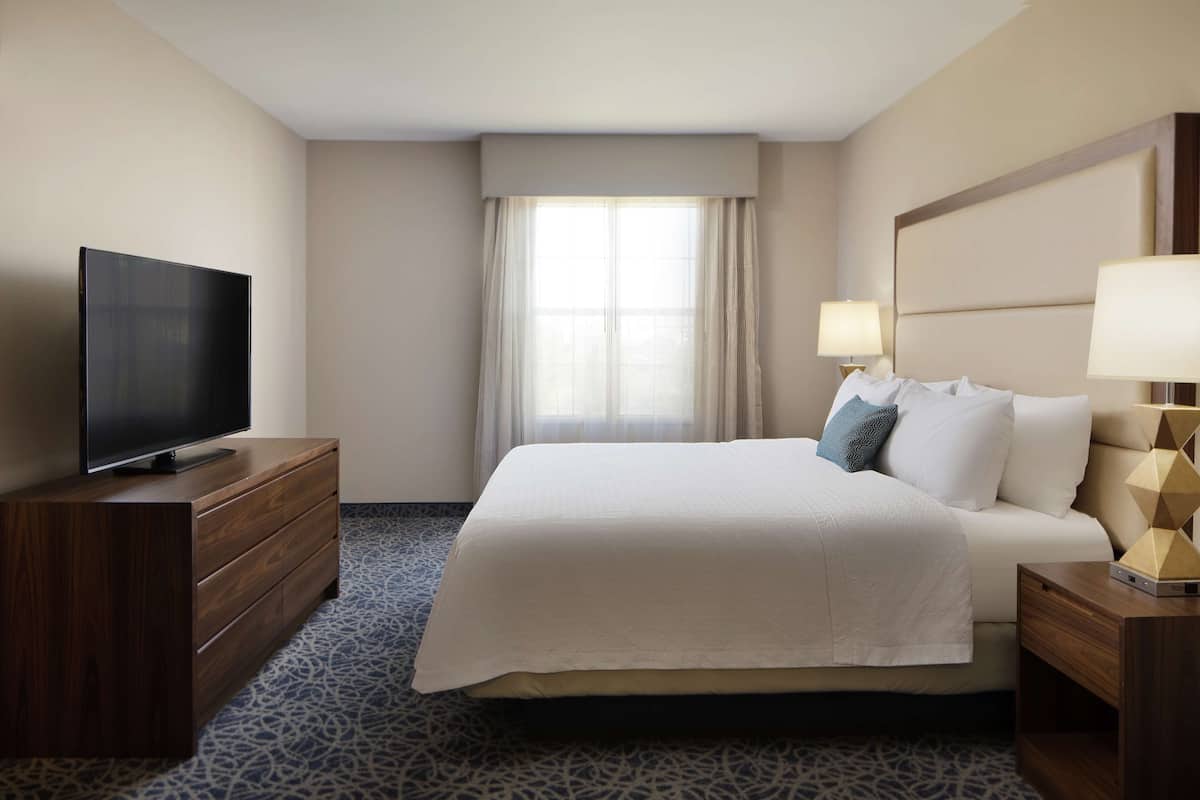 A hotel room with a large bed, a flat-screen TV on a wooden dresser, and a nightstand with a lamp.