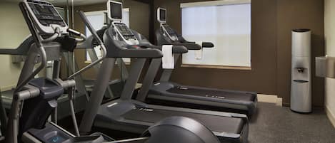 Un gimnasio con cintas de correr y equipo de ejercicio, un espejo grande y una ventana con persianas.