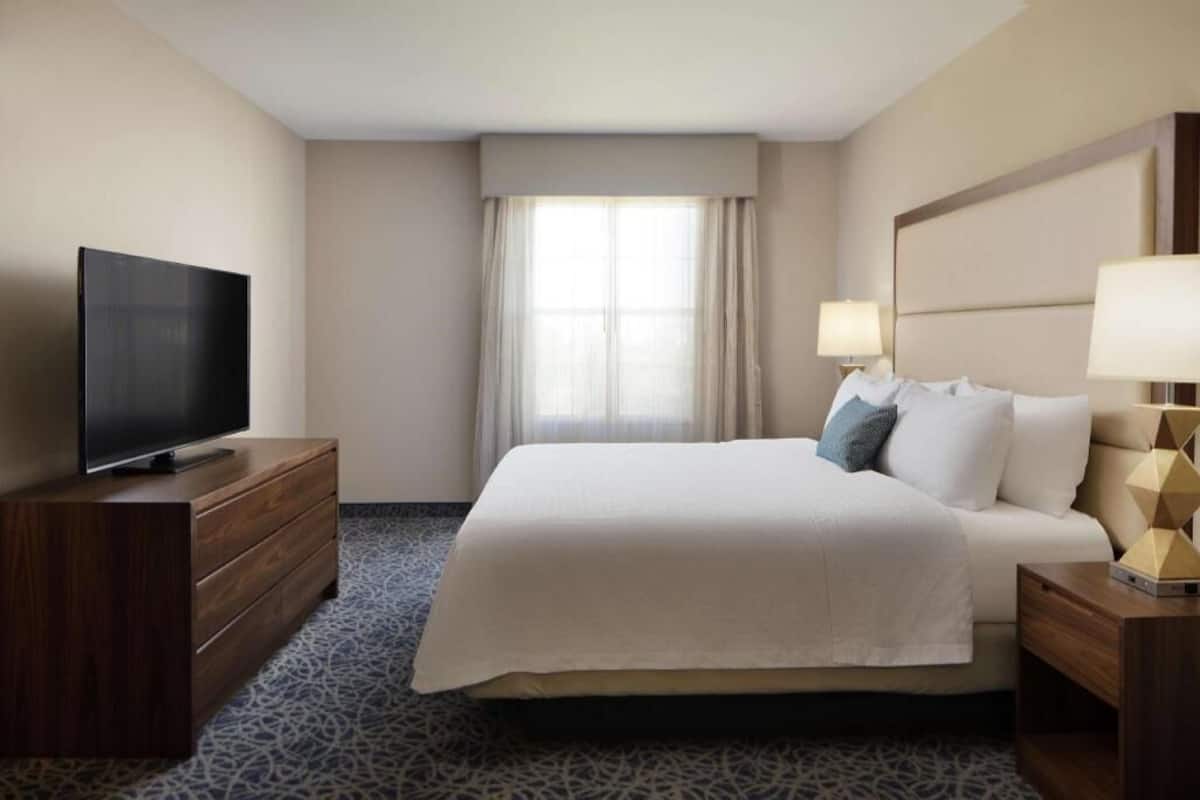 A hotel room with a bed, a television on a wooden dresser, a lamp, and a window with curtains.
