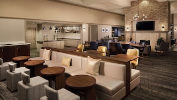 A spacious hotel lobby with a modern design, featuring a large seating area with sofas and chairs, a bar area, and a television mounted on a stone wall.