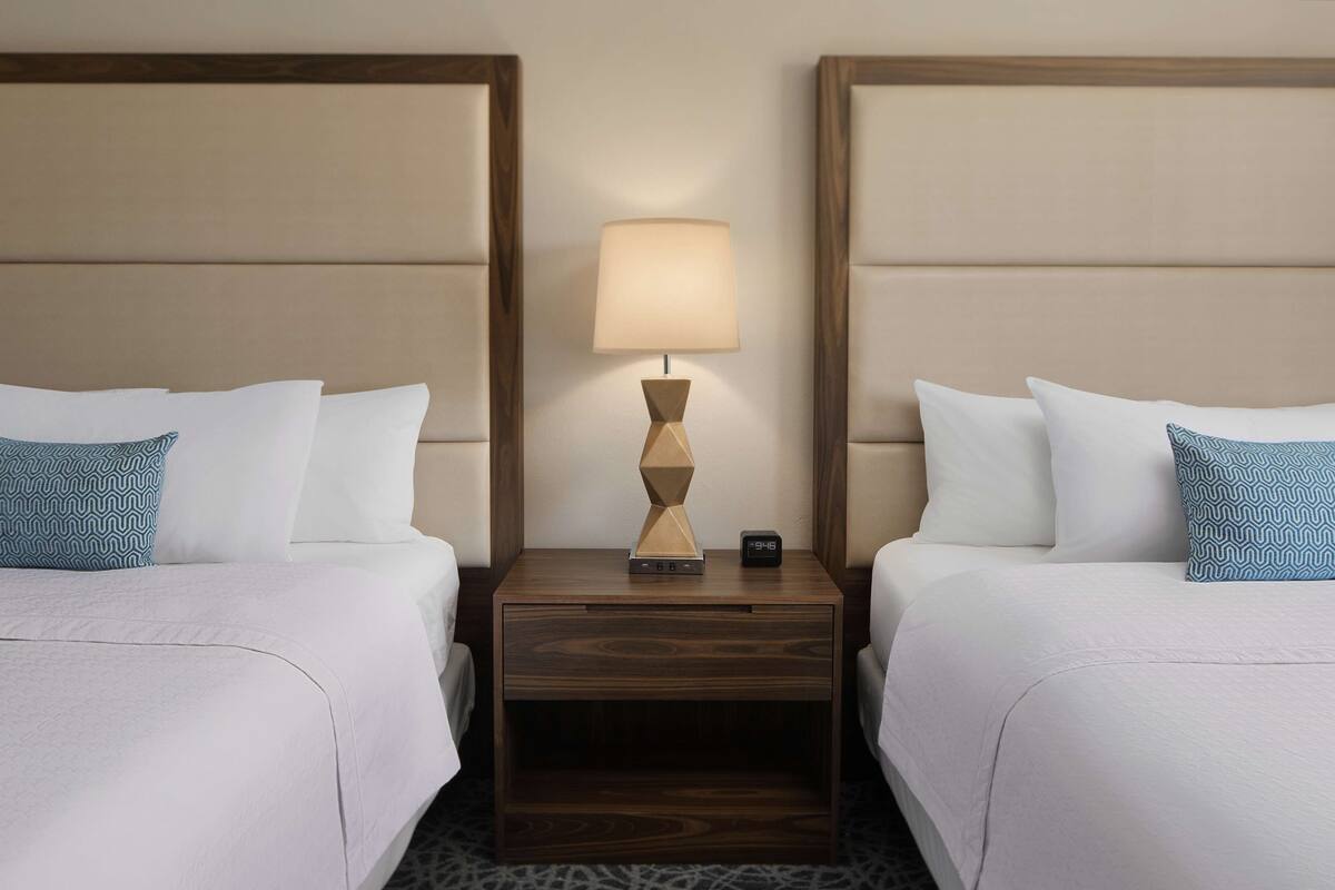 A hotel room with two beds, a nightstand with a lamp, and a digital clock.
