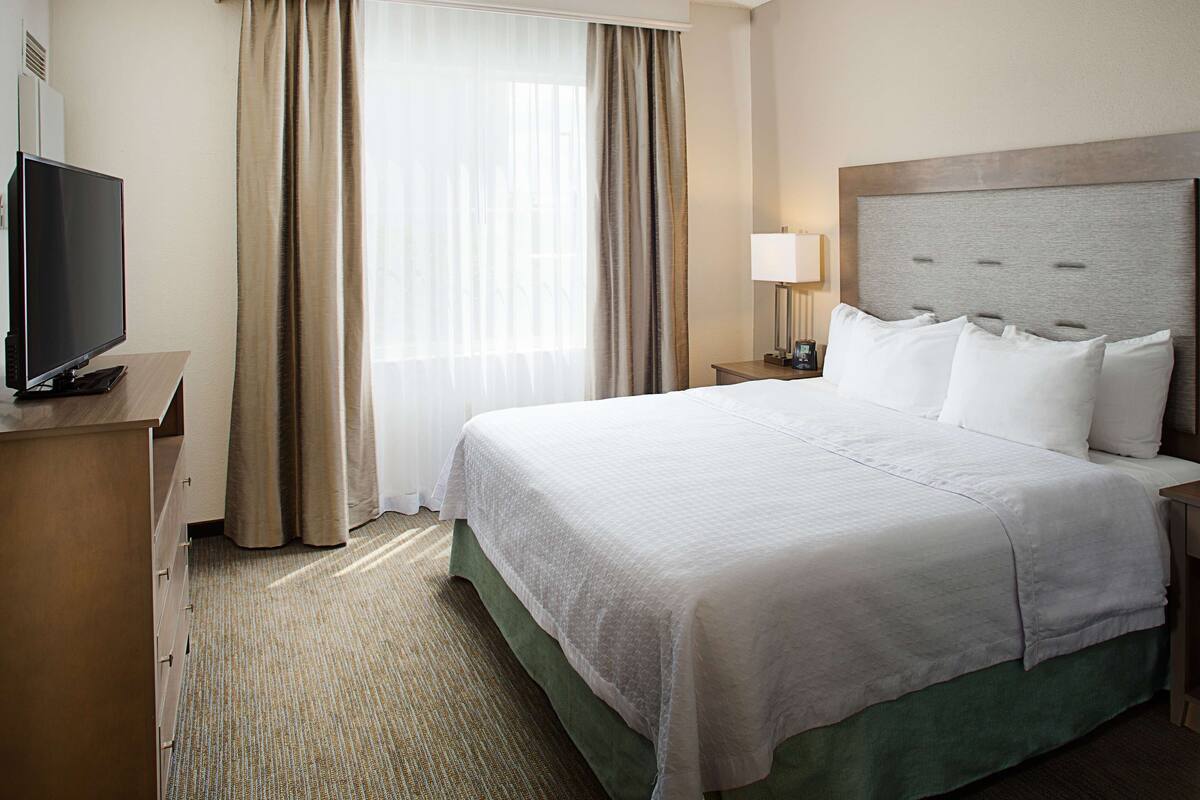 A hotel room with a large bed, a television on a wooden dresser, a nightstand with a lamp, and a window with curtains.