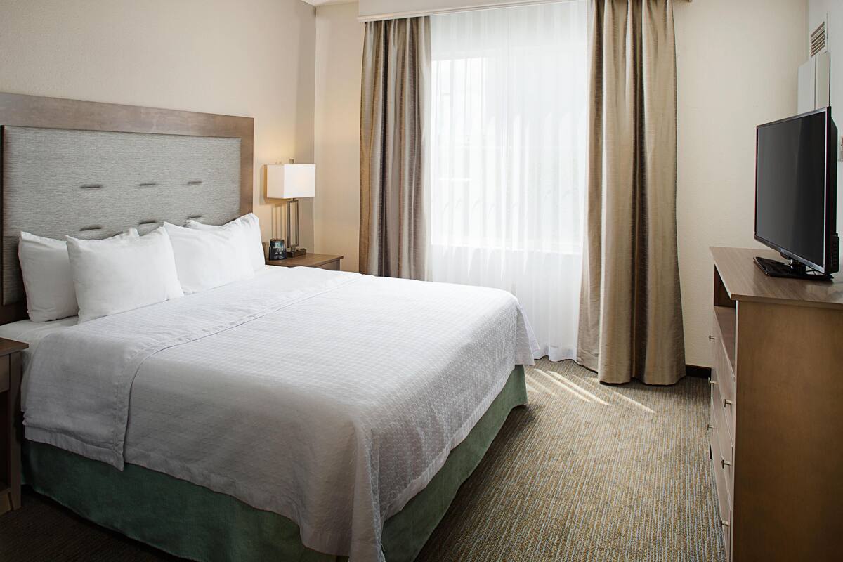 A hotel room with a large bed, a television on a wooden dresser, a nightstand with a lamp, and a window with curtains.